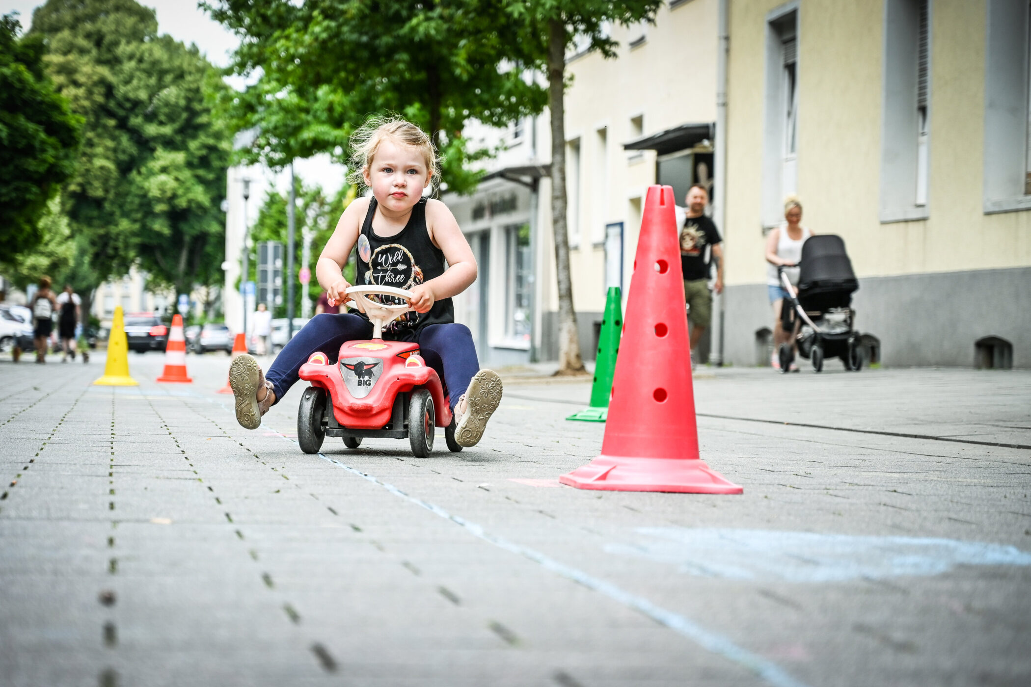 Kind fährt auf rotem Spielzeugauto zwischen bunten Verkehrshütchen auf Gehweg.