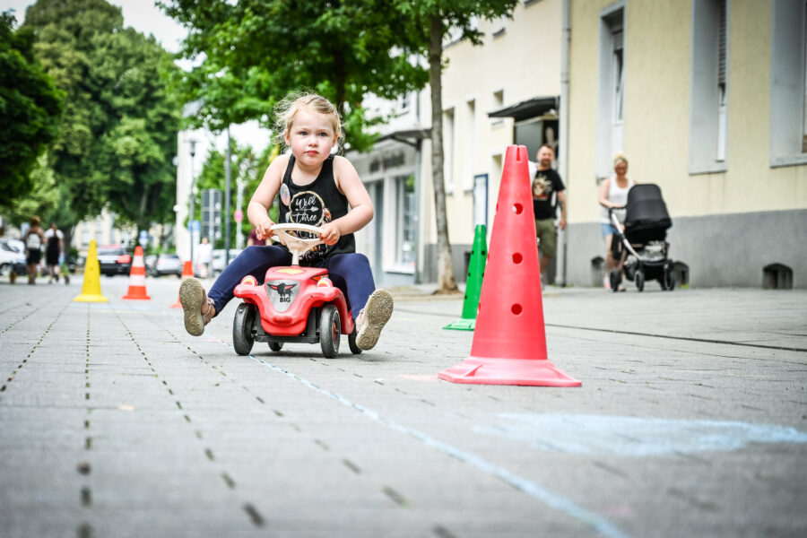 Kind fährt auf rotem Spielzeugauto zwischen bunten Verkehrshütchen auf Gehweg.