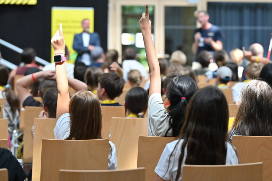 Kinder heben Hände in voller Aula, zwei Erwachsene vorne unscharf.
