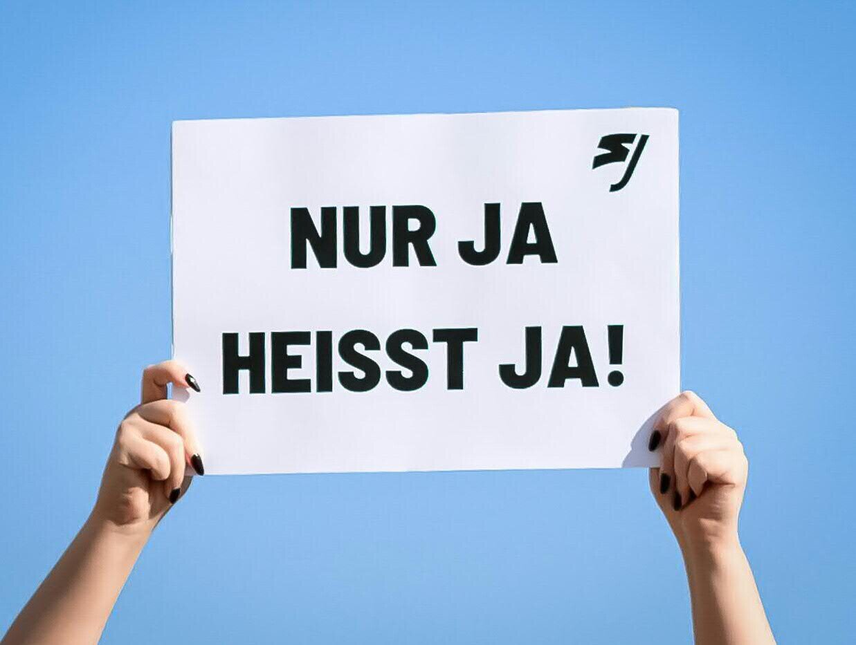 Zwei Hände halten ein Schild mit der Aufschrift "NUR JA HEISST JA!" vor blauem Himmel.