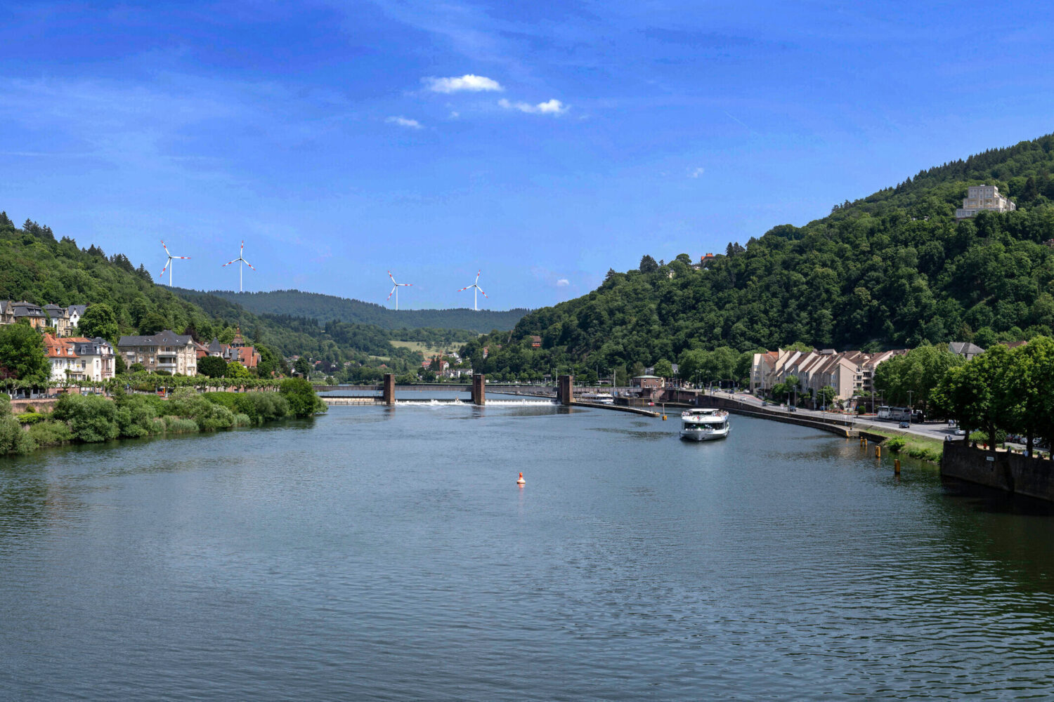 Fluss mit Boot, Brücke, Wohnhäusern und Hügeln mit Windrädern im Hintergrund.