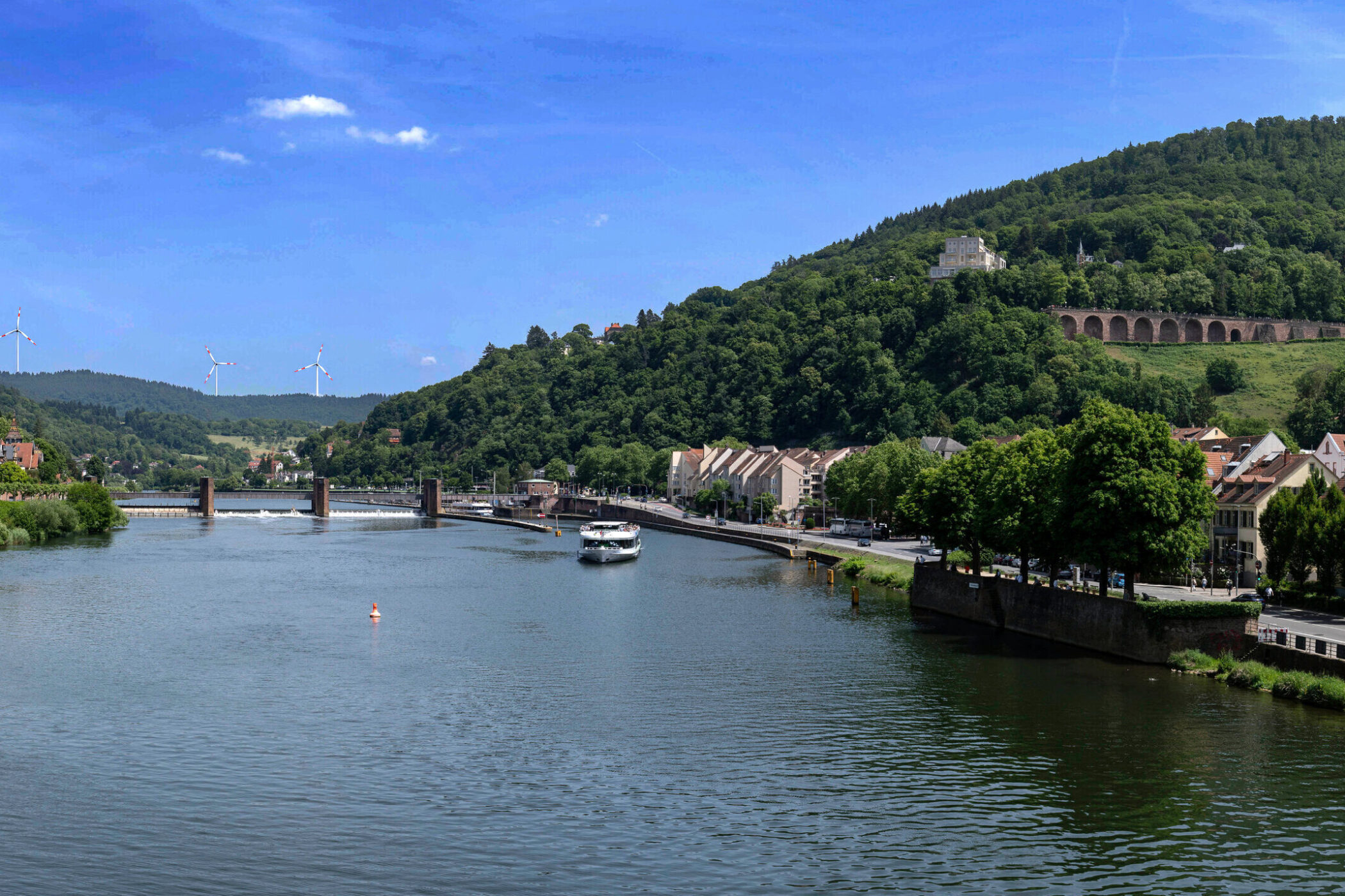 Fluss mit Boot, Brücke, Häusern und waldigen Hügeln im Hintergrund.