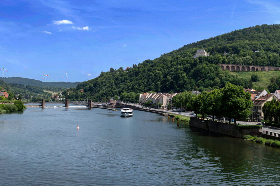 Fluss mit Boot, Brücke, Häusern und waldigen Hügeln im Hintergrund.