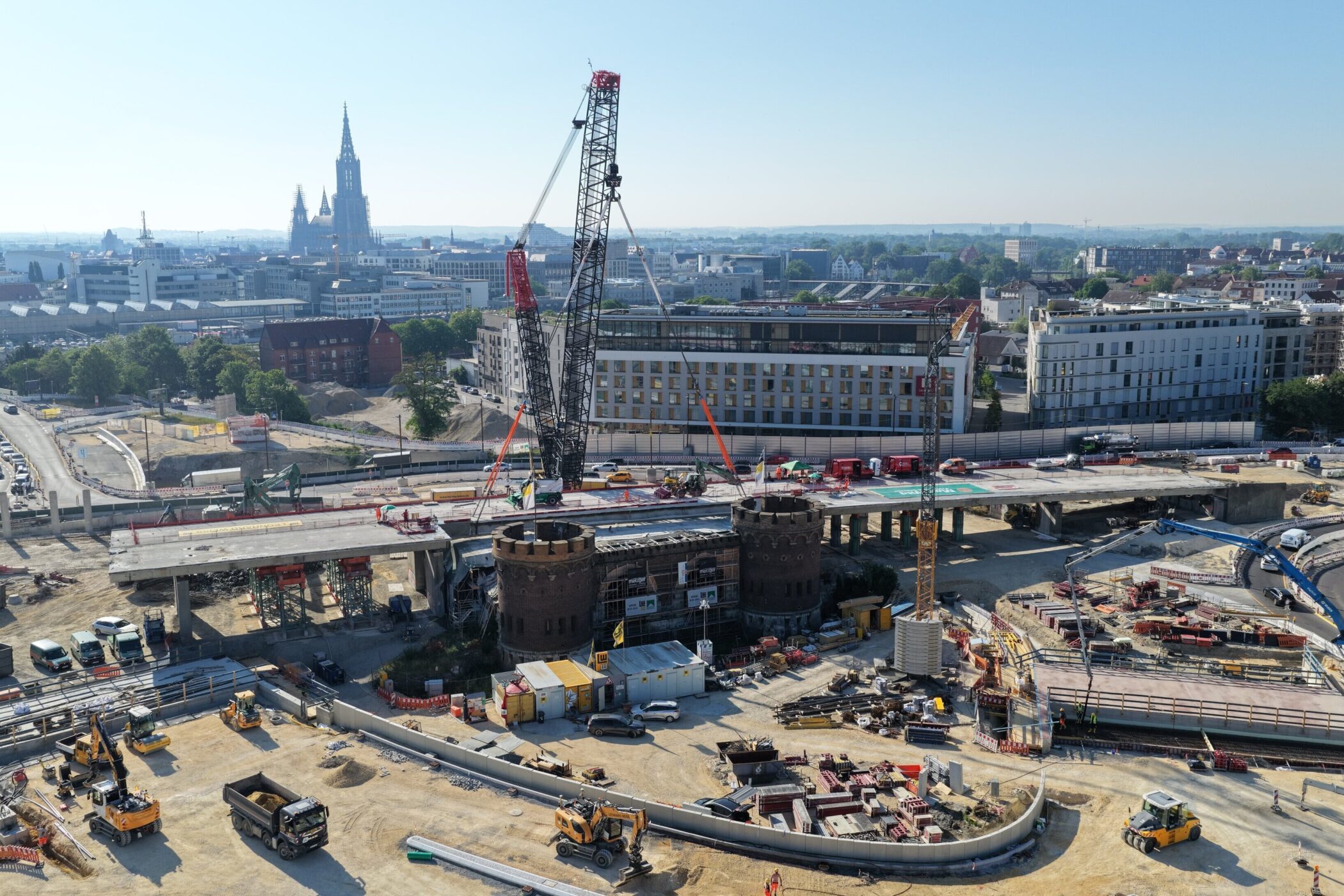 Baustelle mit Kran, LKW, Bagger und mehreren Gebäuden im Hintergrund.