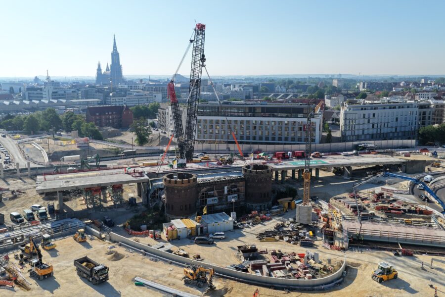 Baustelle mit Kran, LKW, Bagger und mehreren Gebäuden im Hintergrund.