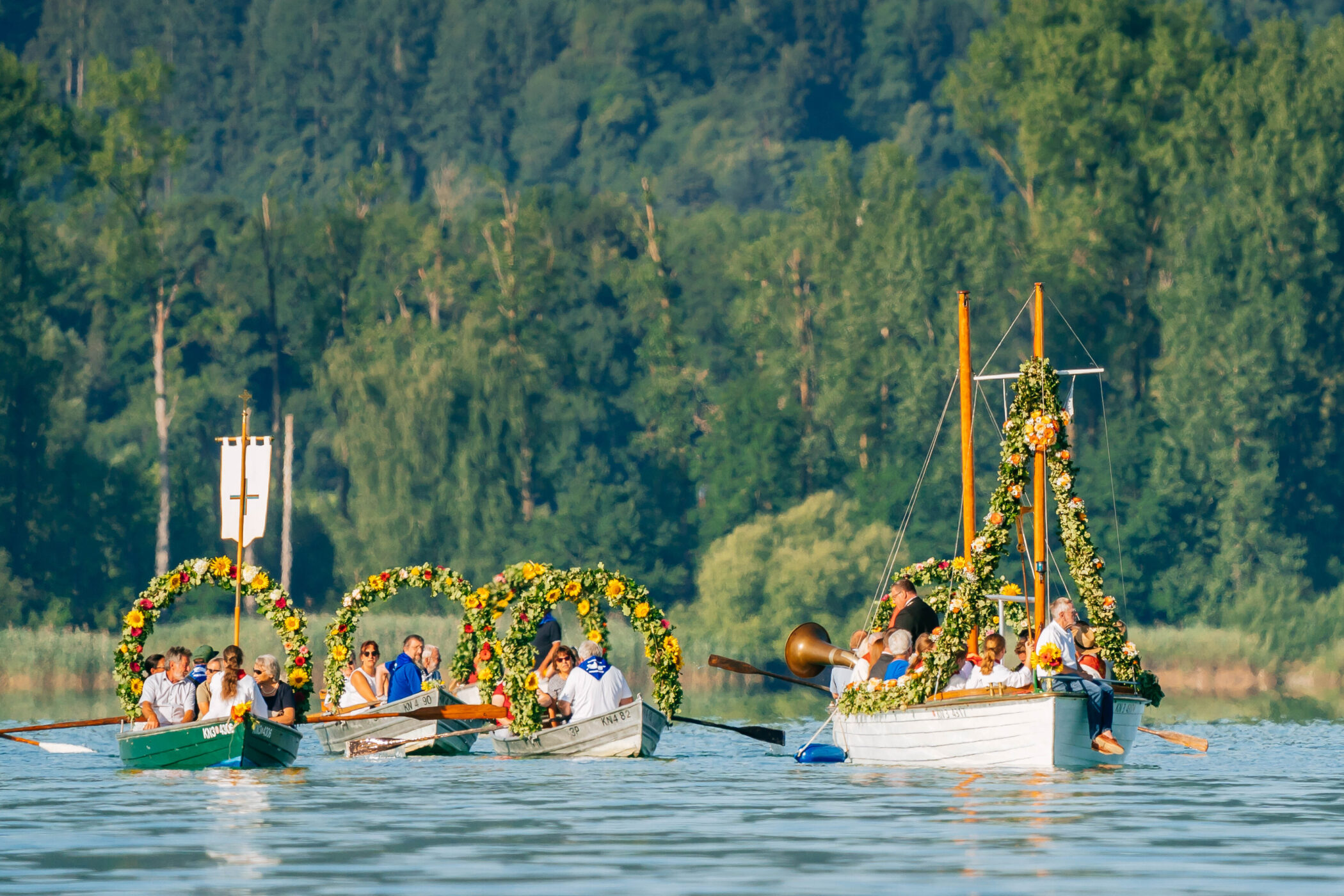 Mehrere Boote mit Blumen geschmückt, Personen auf einem See in bewaldeter Umgebung.