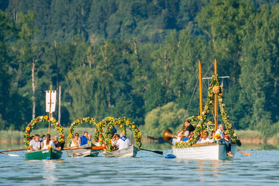 Mehrere Boote mit Blumen geschmückt, Personen auf einem See in bewaldeter Umgebung.