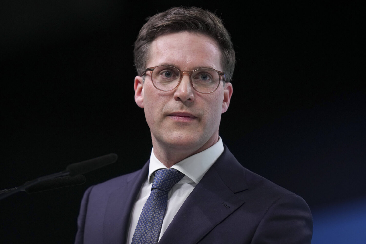 Eine Person mit Brille spricht vor einem Mikrofon, trägt Anzug und Krawatte.