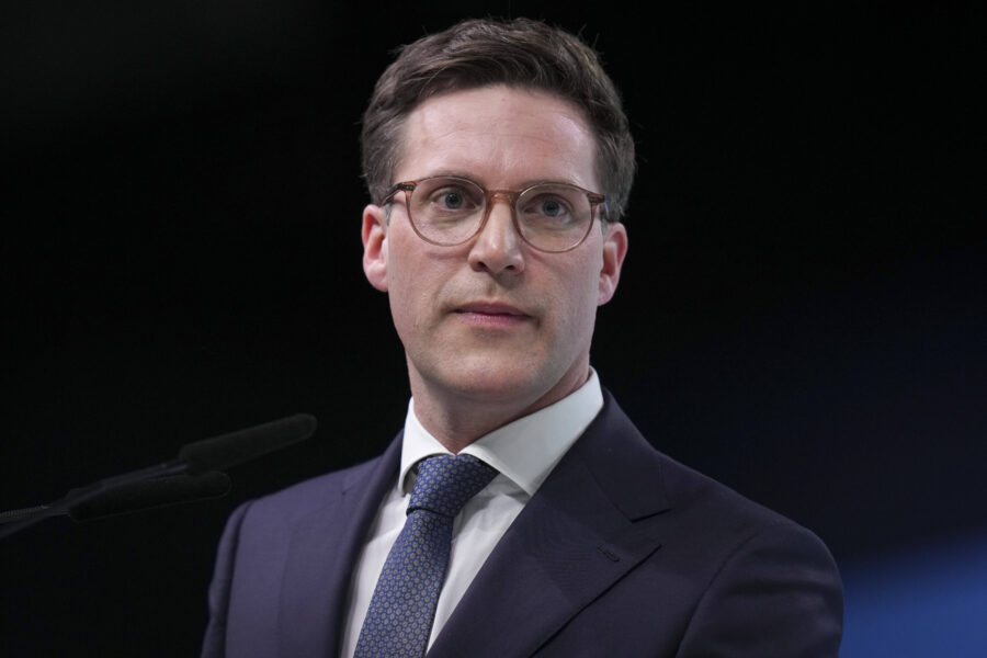 Eine Person mit Brille spricht vor einem Mikrofon, trägt Anzug und Krawatte.