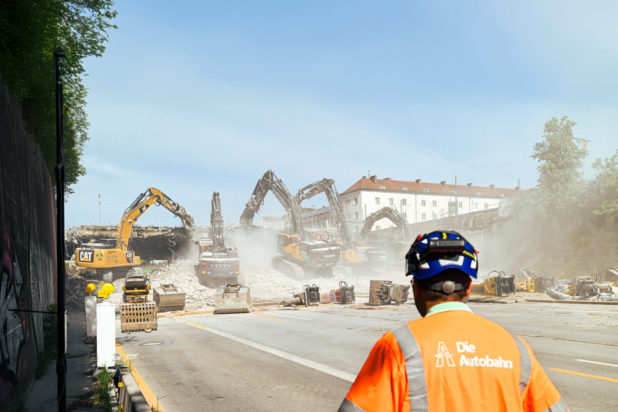 Mann in orangefarbener Weste beobachtet Bagger beim Abriss einer Brücke.