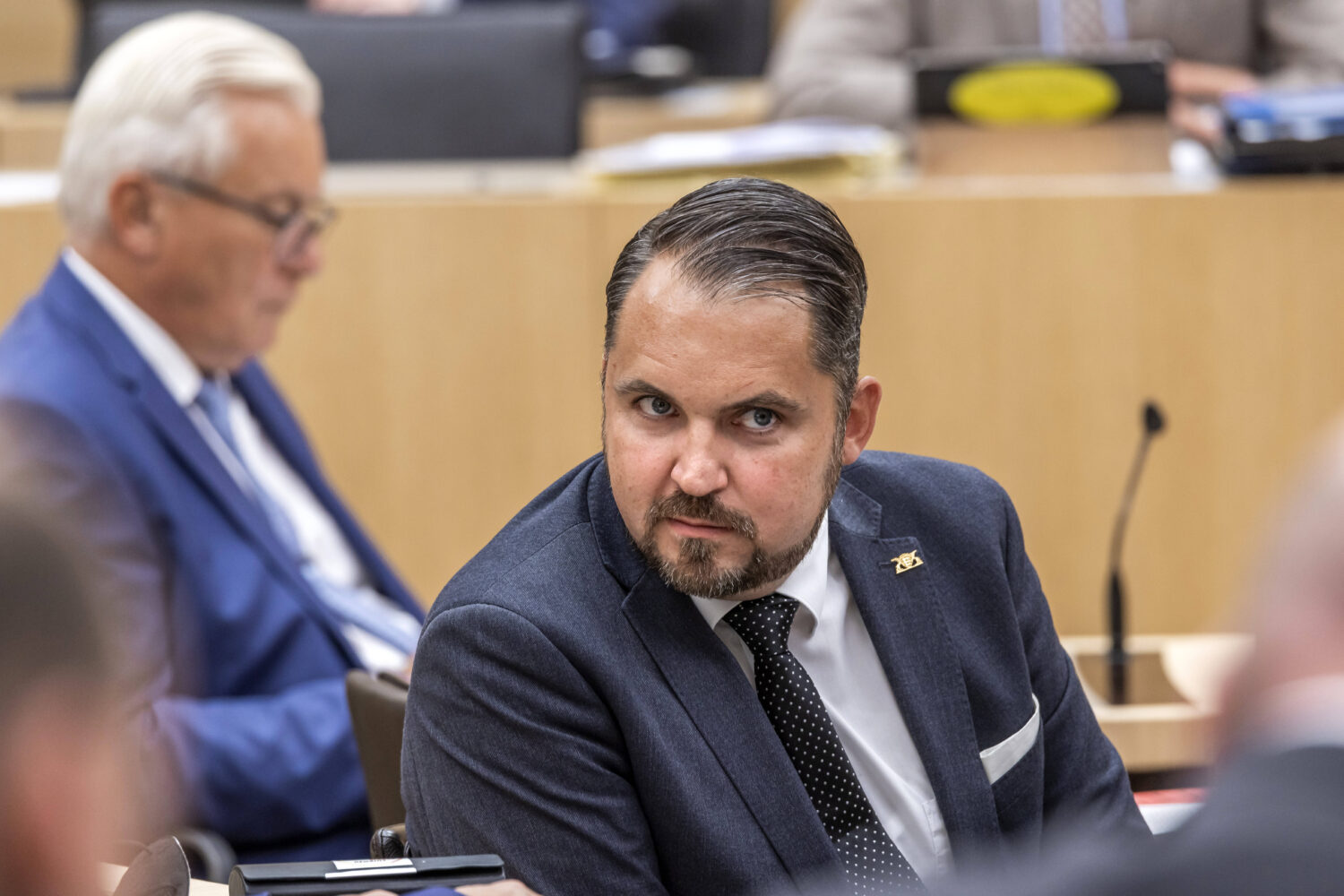 Mann in Anzug sitzt in einem Raum, weitere Personen im Hintergrund.