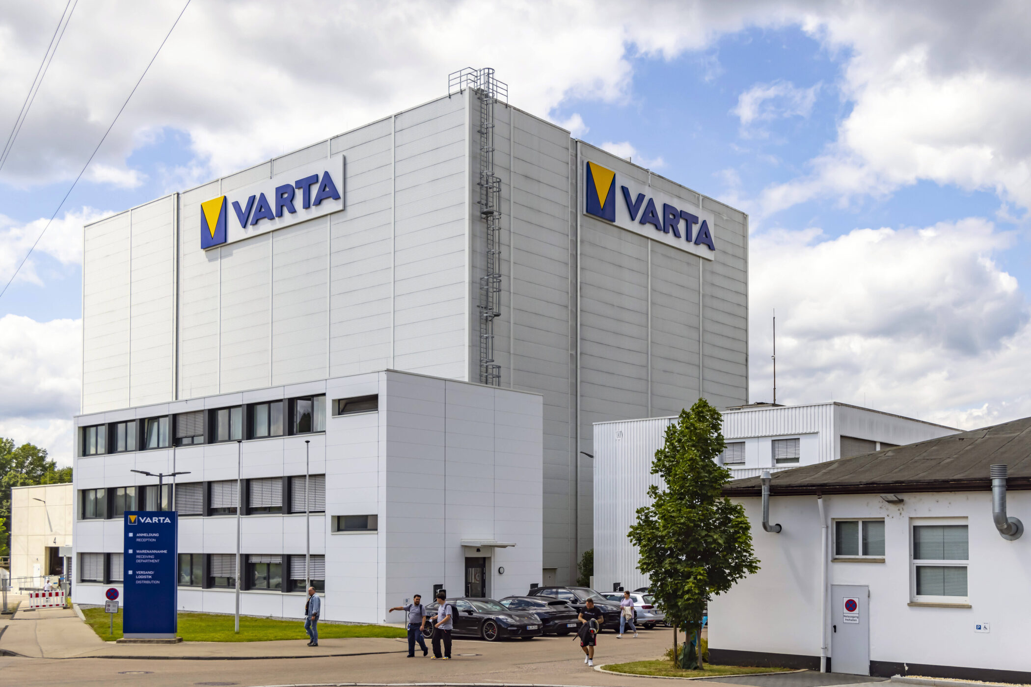 Großes Gebäude mit "VARTA"-Logos, Menschen und Autos davor, blauer Himmel.