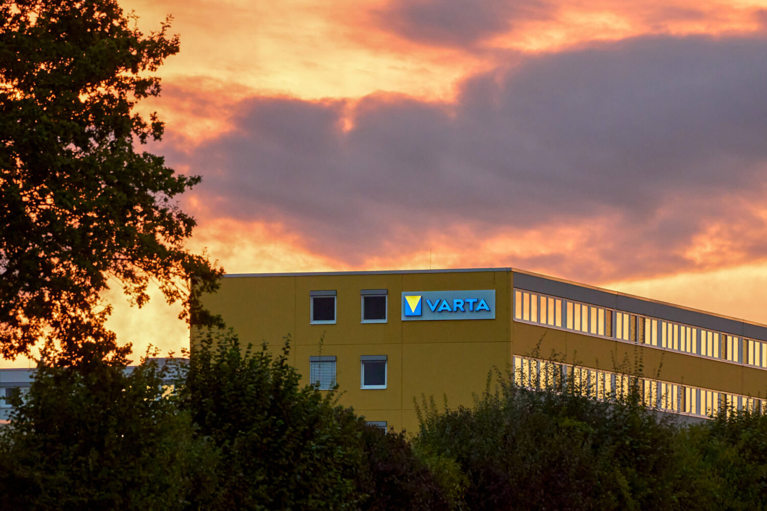 Gebäude mit "VARTA"-Logo vor buntem Abendhimmel, Bäume im Vordergrund.
