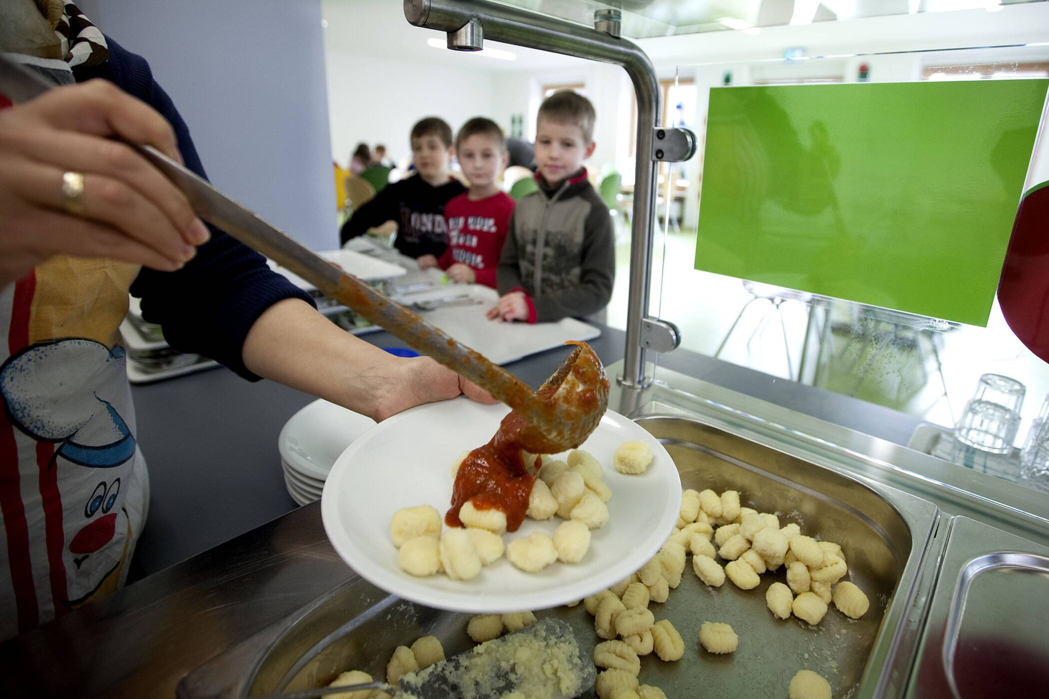 Person schöpft Soße über Gnocchi auf einem Teller, Kinder warten in der Schlange.
