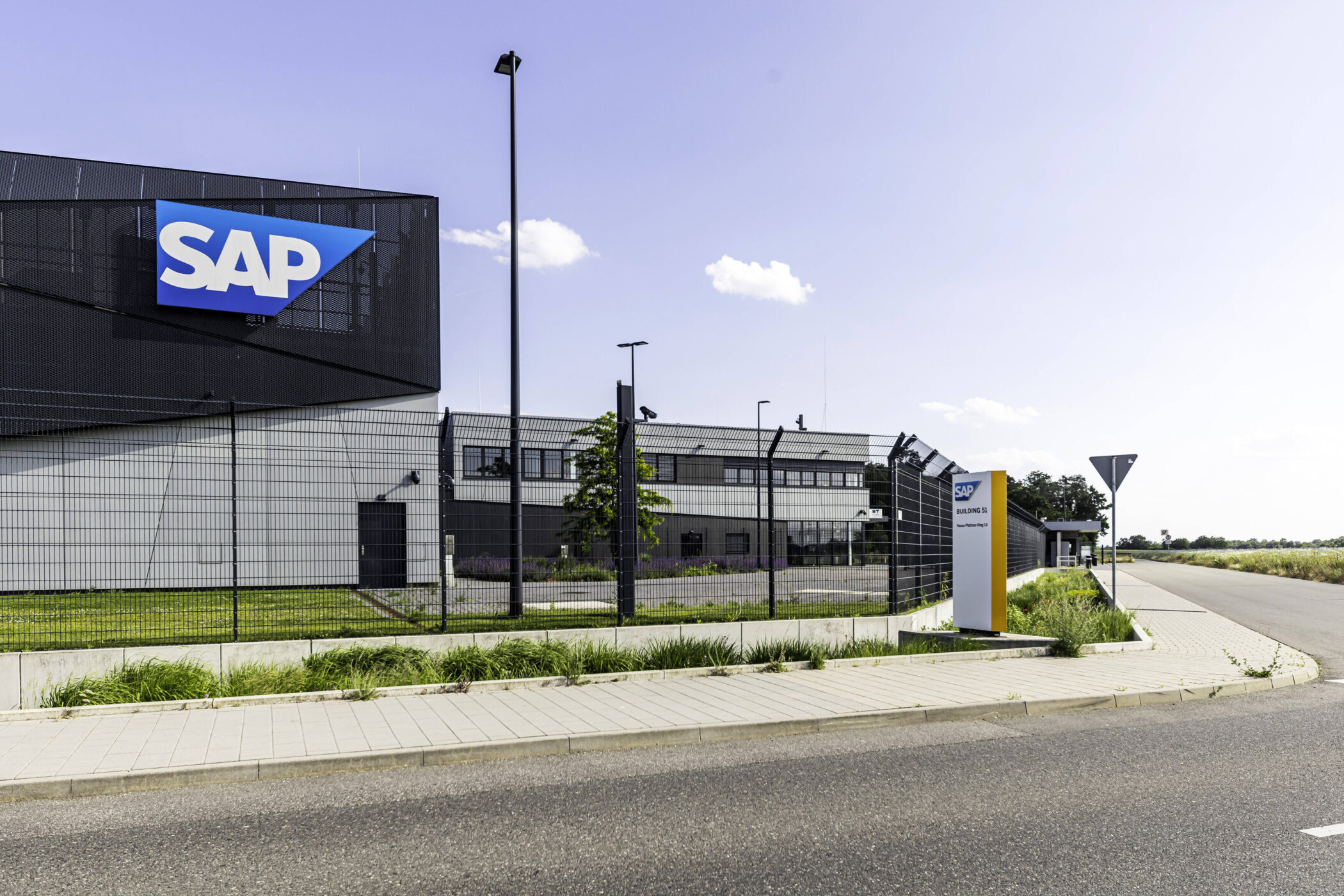 Gebäude mit SAP-Logo, umzäunt, davor Straße und Gehweg.