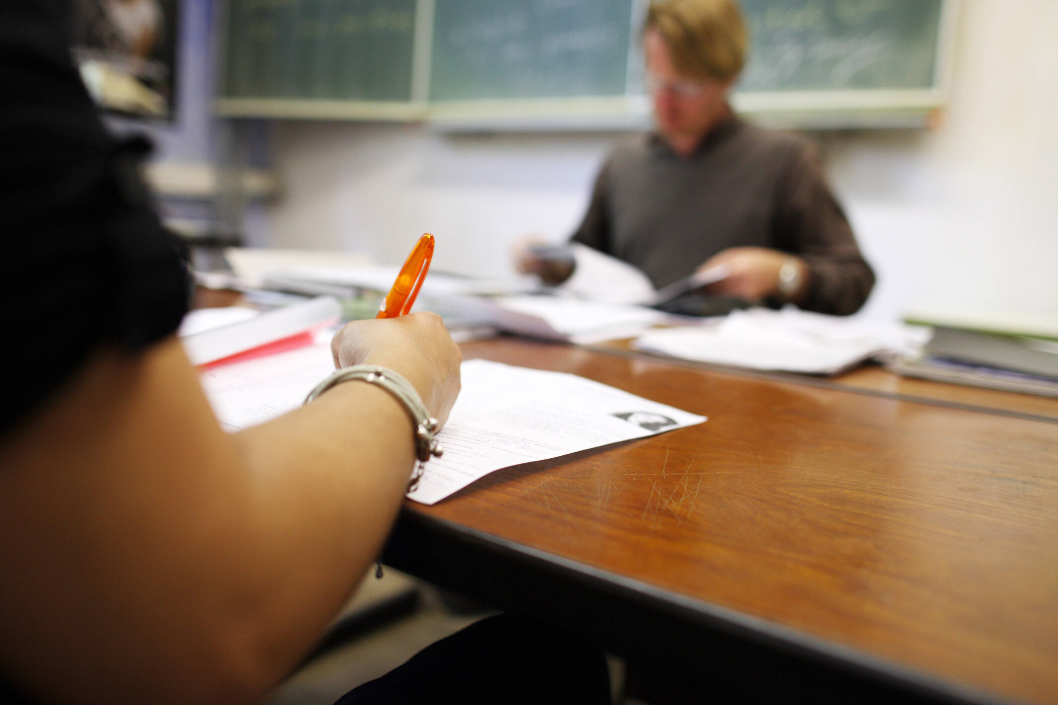 Person schreibt mit orangem Stift an Tisch, zweite Person unscharf im Hintergrund mit Papier.