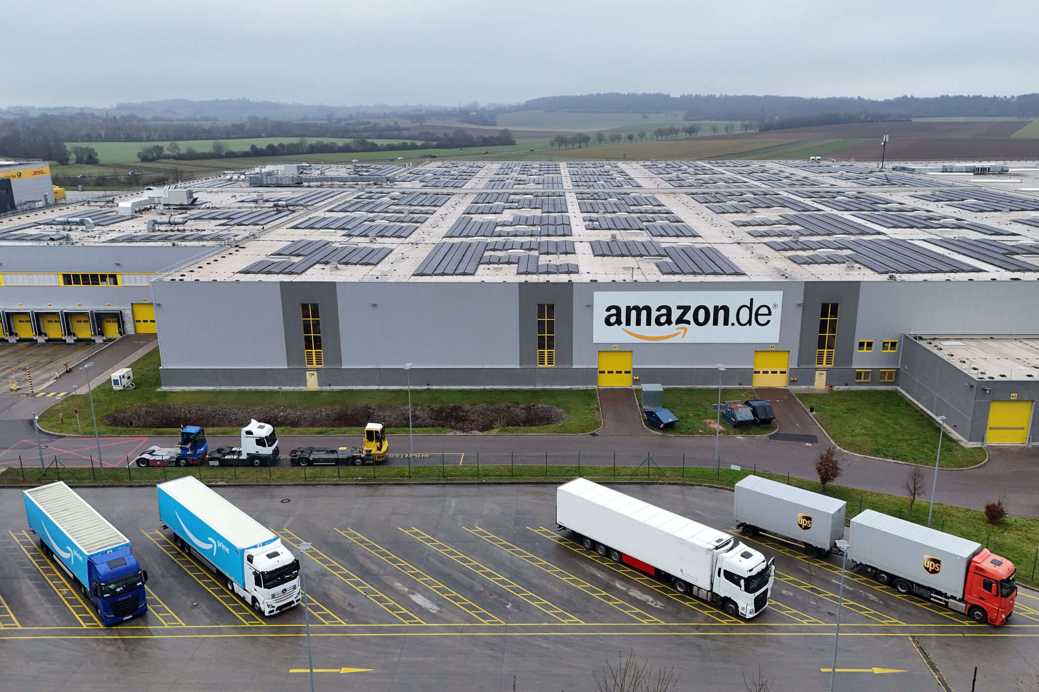 Verteilzentrum mit LKWs, großes amazon.de-Logo an grauem Gebäude, Solarpaneele auf Dach.
