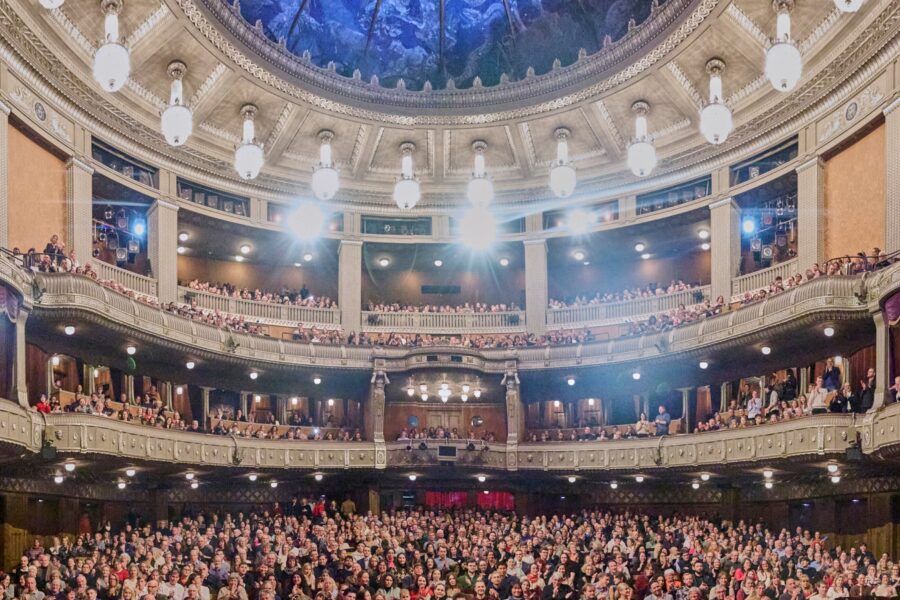 Volles Theaterpublikum, mehrere Ränge, große Deckenlichter.