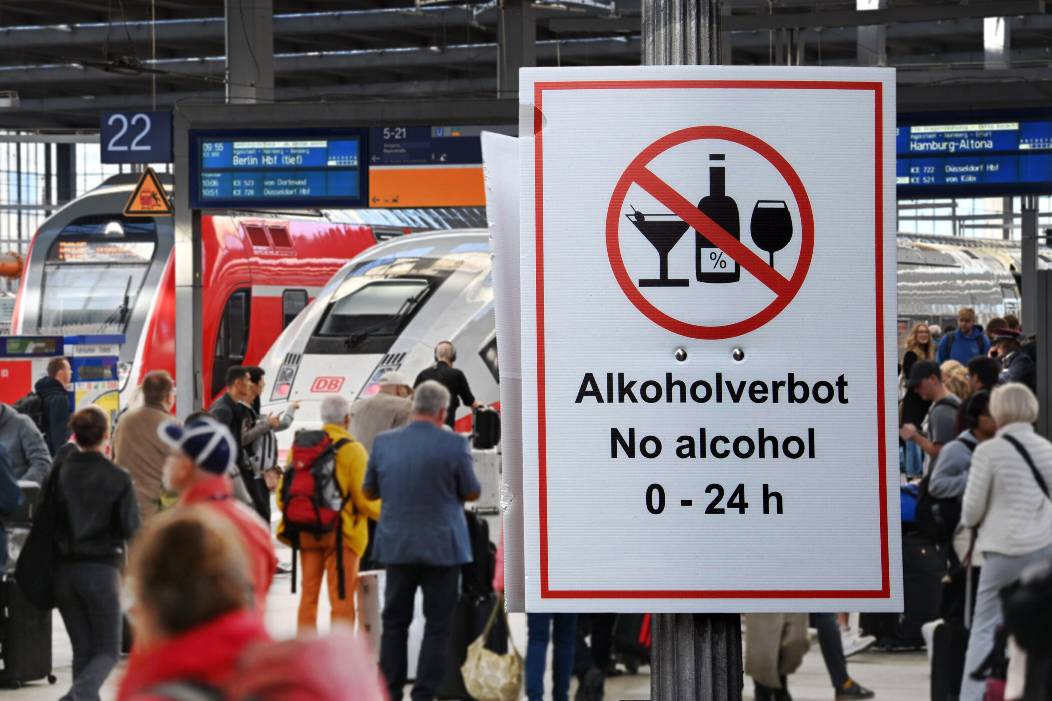 Schild mit Alkoholverbot, Menschen und Züge im Hintergrund am Bahnsteig.