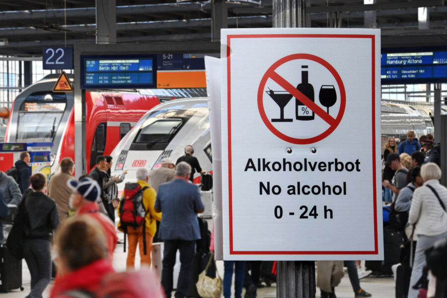 Schild mit Alkoholverbot, Menschen und Züge im Hintergrund am Bahnsteig.