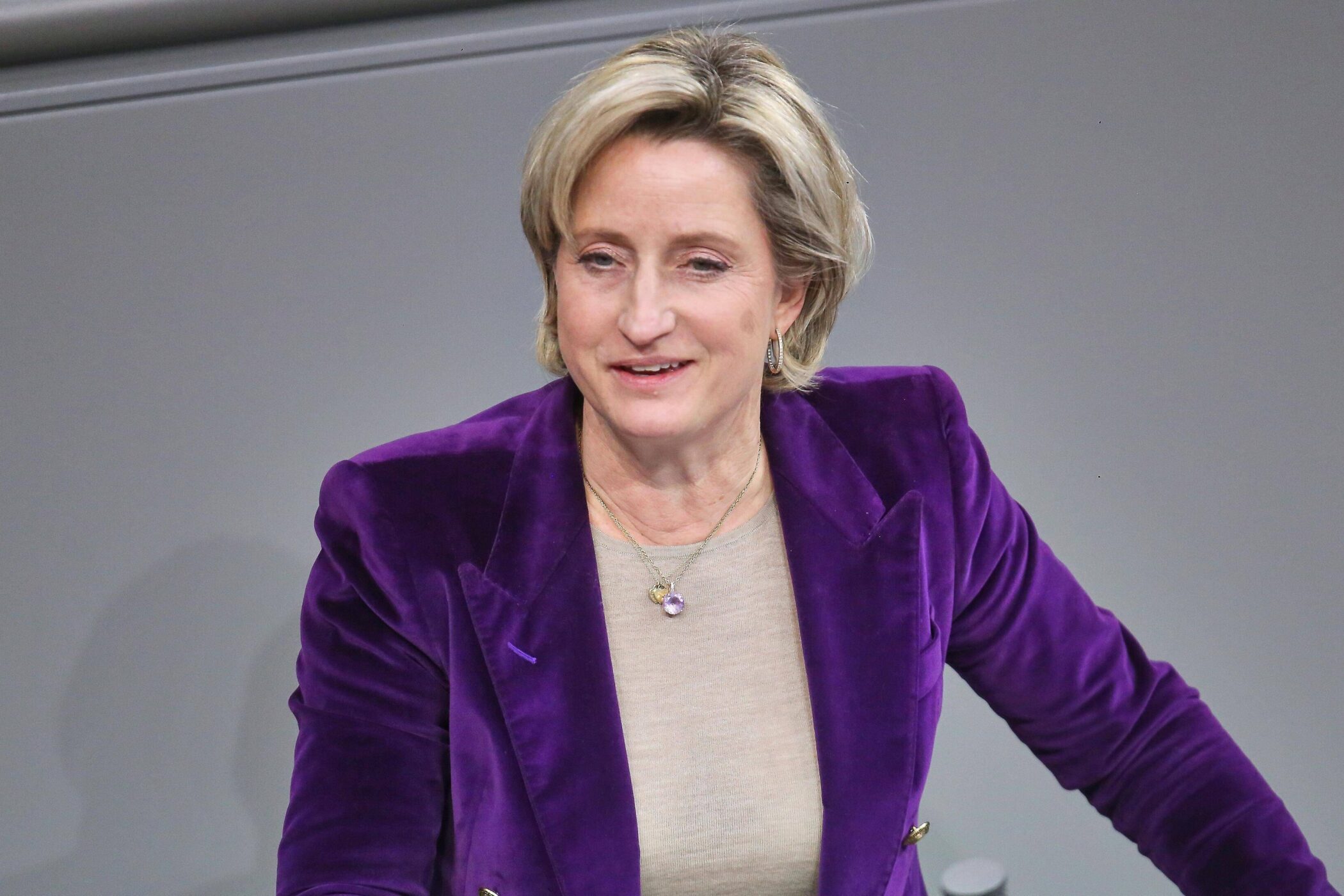 Person mit lila Blazer spricht, hellgrauer Hintergrund.