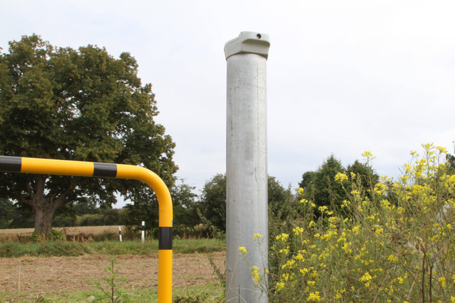 Metallmast neben gelbem Stahlrohr, Bäume und gelbe Blumen im Hintergrund.