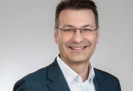 Mann mit Brille und Anzug, lächelnd vor grauem Hintergrund.
