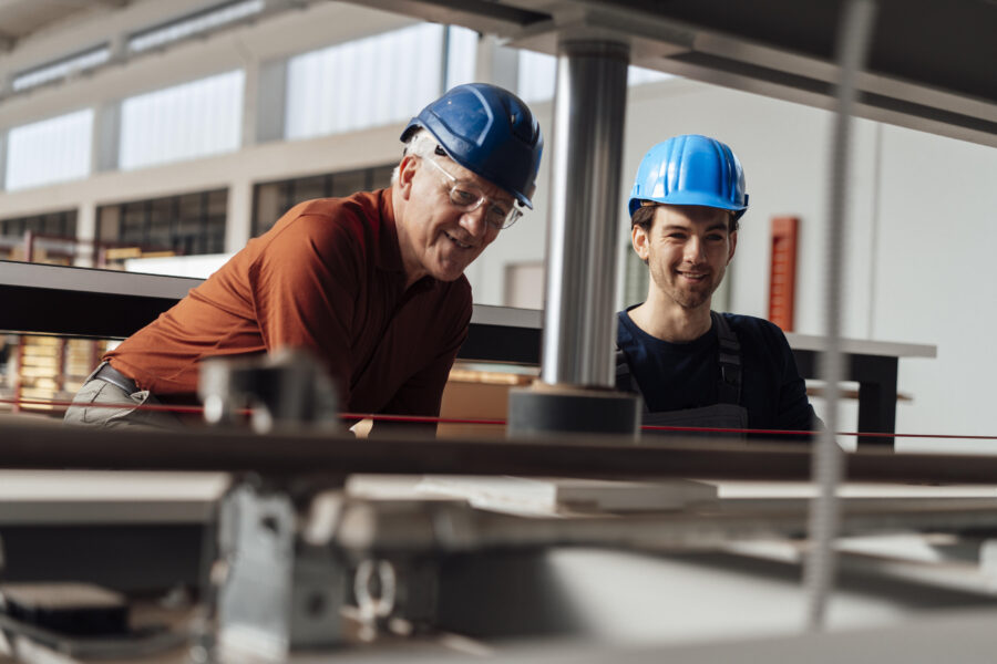 Zwei Personen mit blauen Schutzhelmen arbeiten in einer Fabrikumgebung.