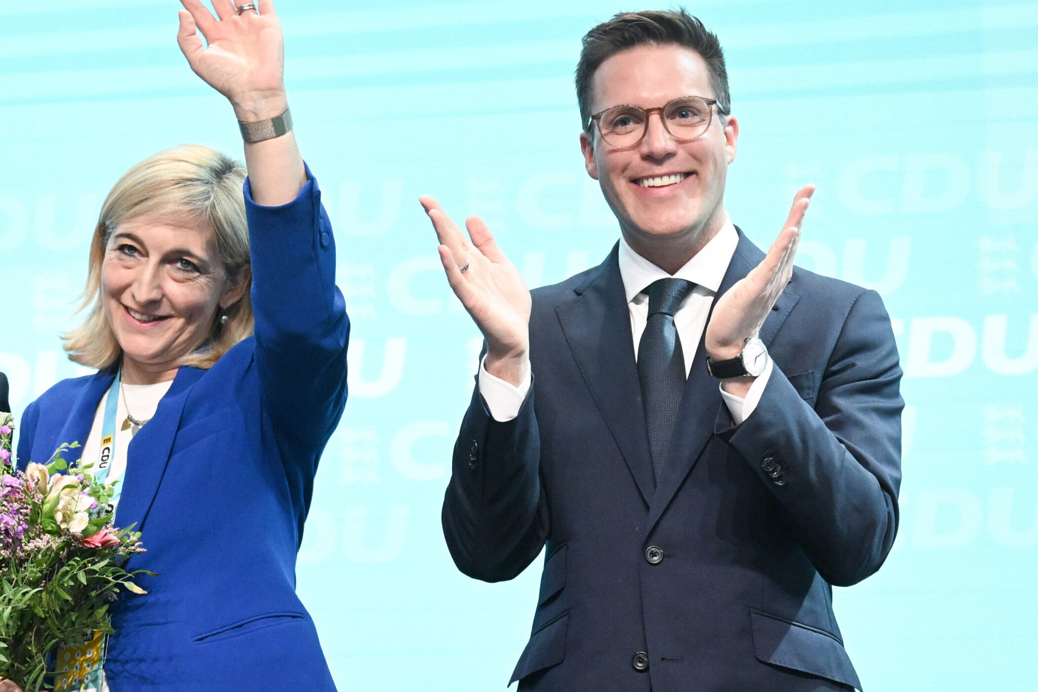 Zwei Personen, eine winkend mit Blumen, der andere klatschend, vor CDU-Hintergrund.