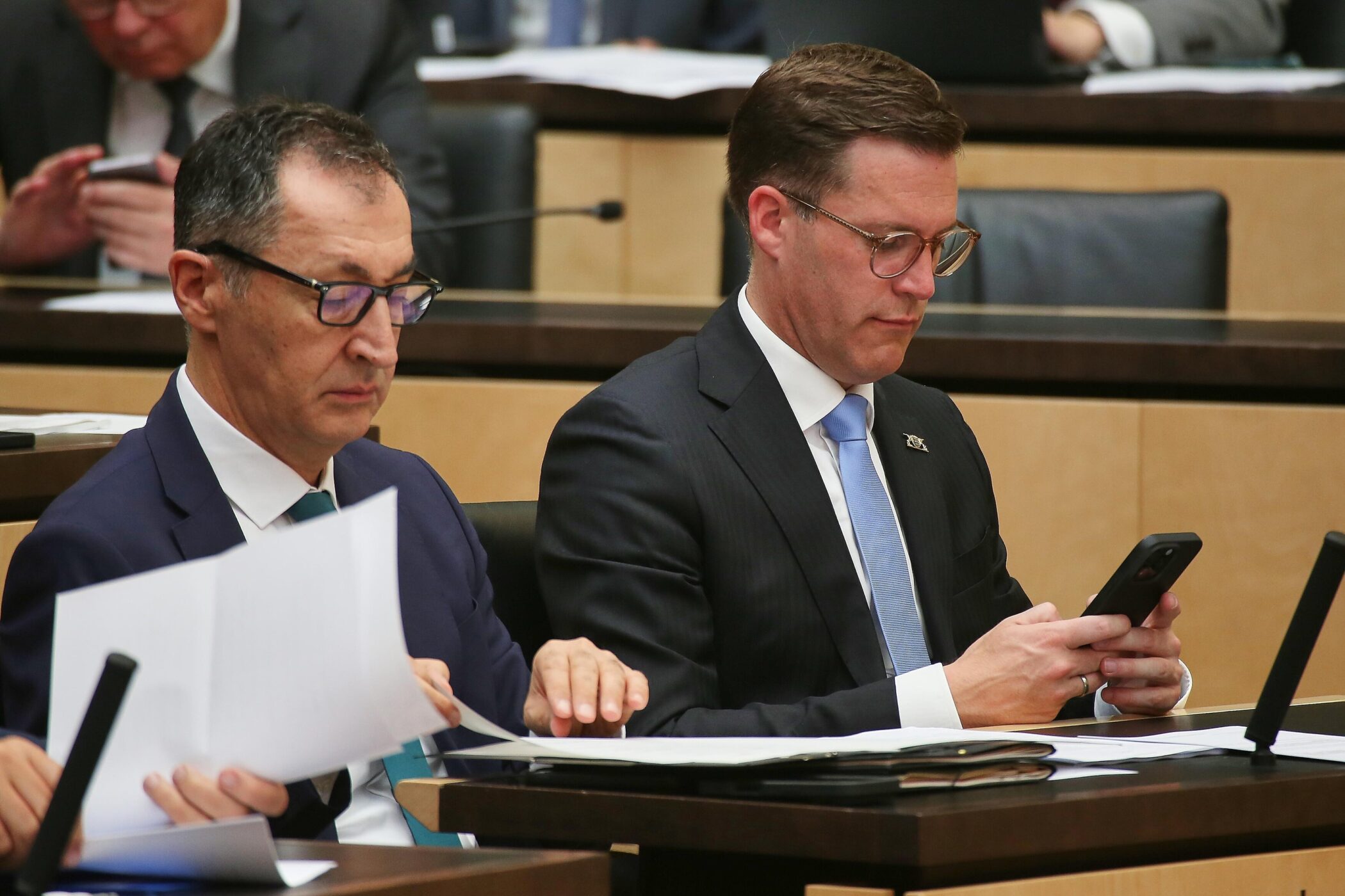 Zwei Männer im Anzug sitzen an einem Tisch, einer hält ein Smartphone.