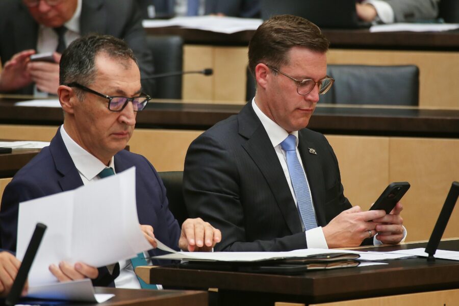 Zwei Männer im Anzug sitzen an einem Tisch, einer hält ein Smartphone.