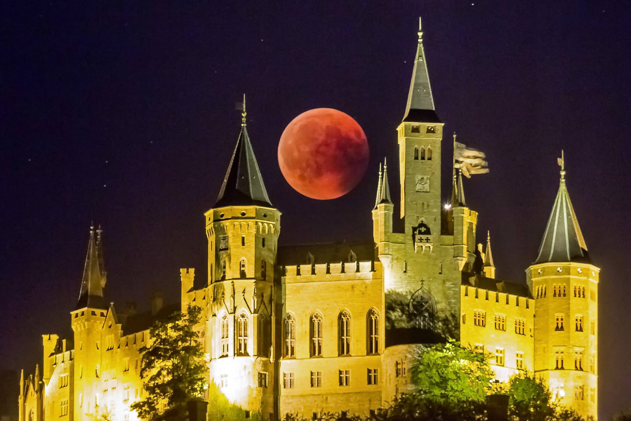 Beleuchtetes Schloss bei Nacht, großer roter Mond am Himmel.