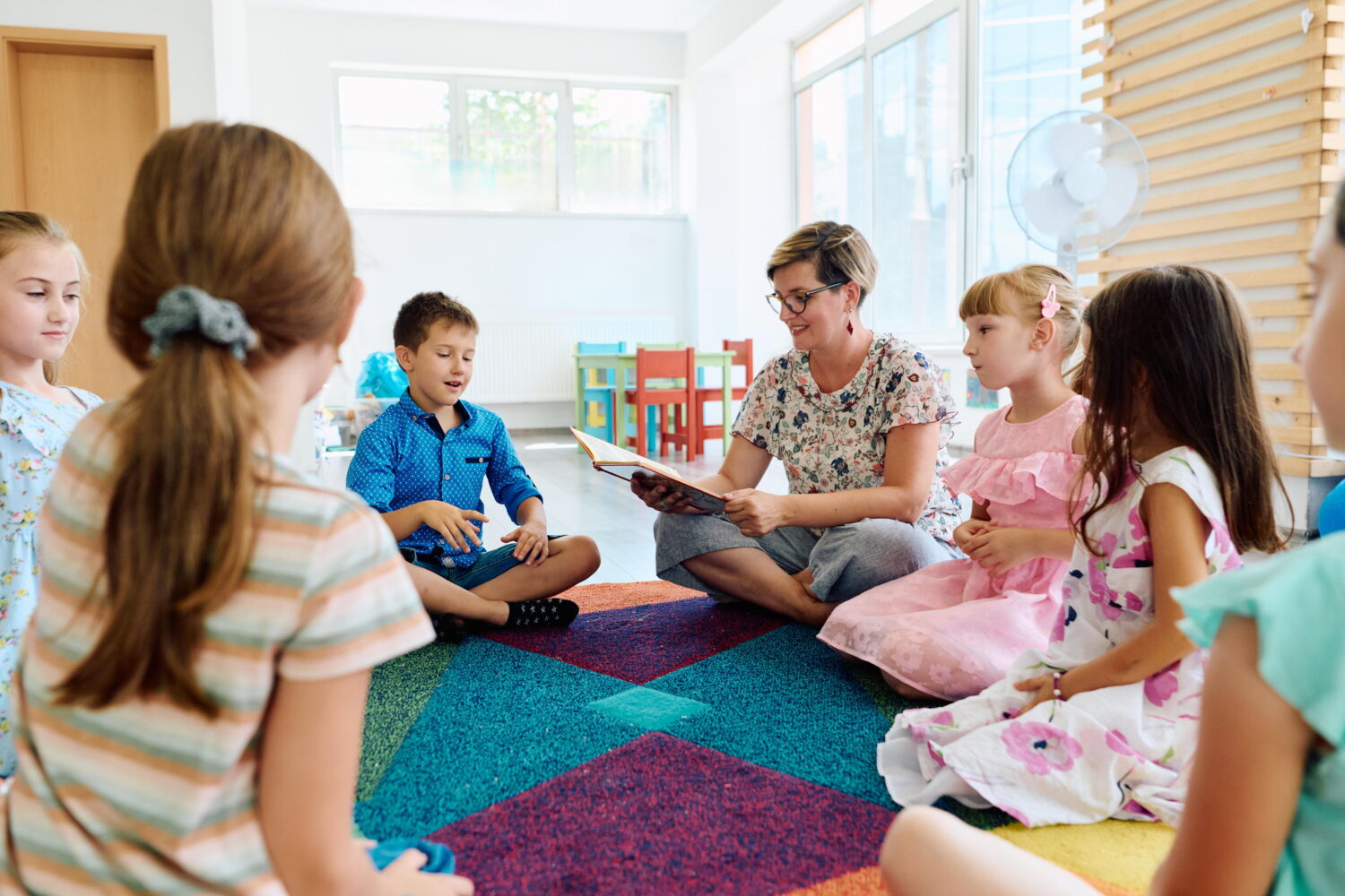 Frau liest einer Gruppe von fünf Kindern im Kreis sitzend ein Buch vor.
