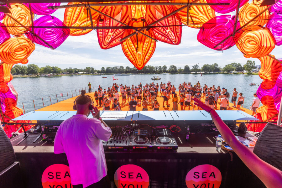 DJ mit Kopfhörern, Menschenmenge tanzt auf orange Plattform am See. Bunte Deko oben.