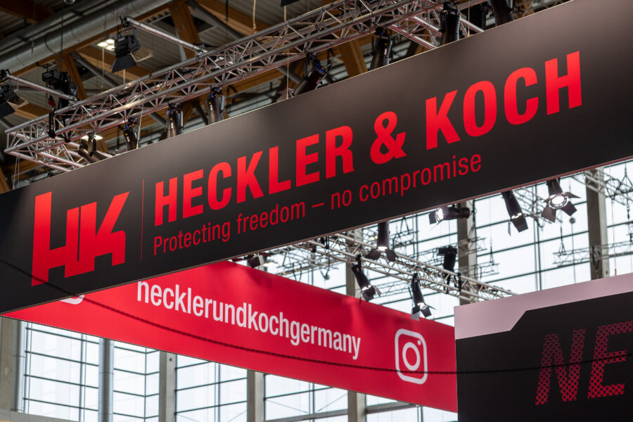 Schwarzes Schild mit rotem "HK" und Text: Heckler & Koch, Protecting freedom.