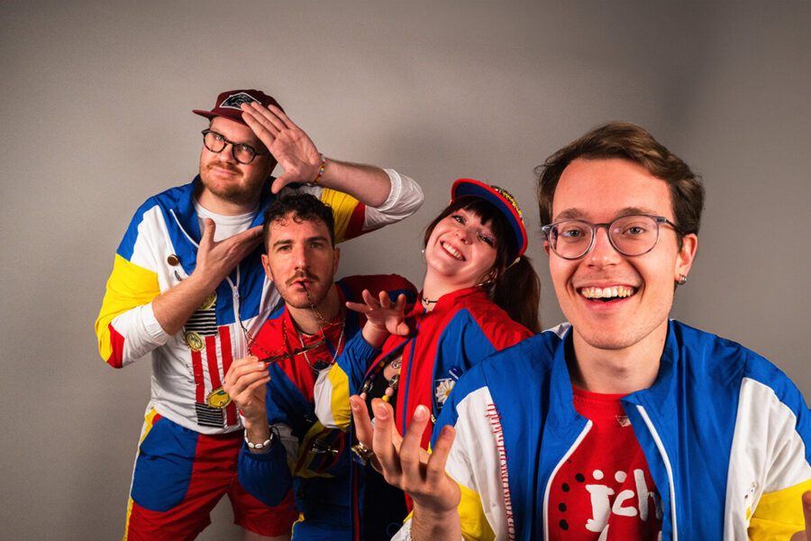 Vier Personen in bunten Outfits posieren fröhlich vor grauem Hintergrund.