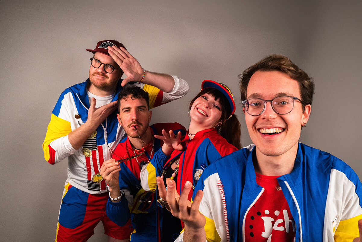 Vier Personen in bunten Outfits posieren fröhlich vor grauem Hintergrund.