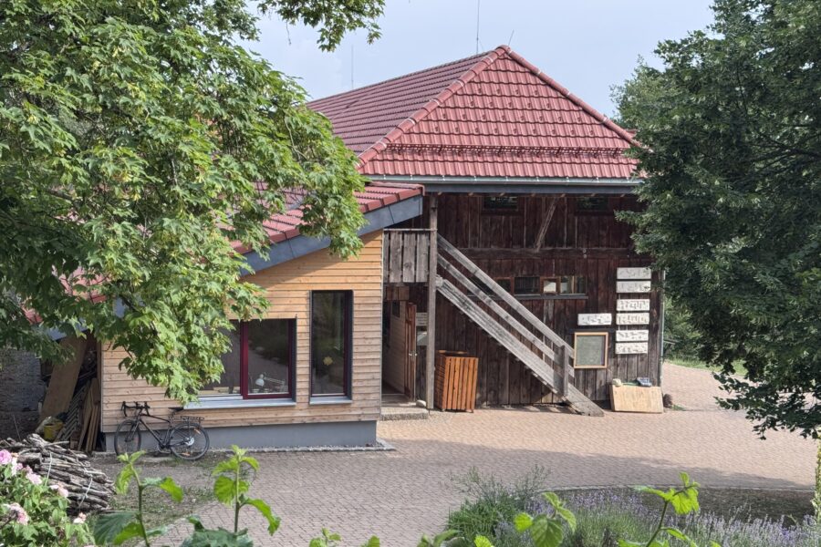 Holzgebäude mit rotem Dach, Fahrrad davor, Bäume und Pflastersteine im Vordergrund.