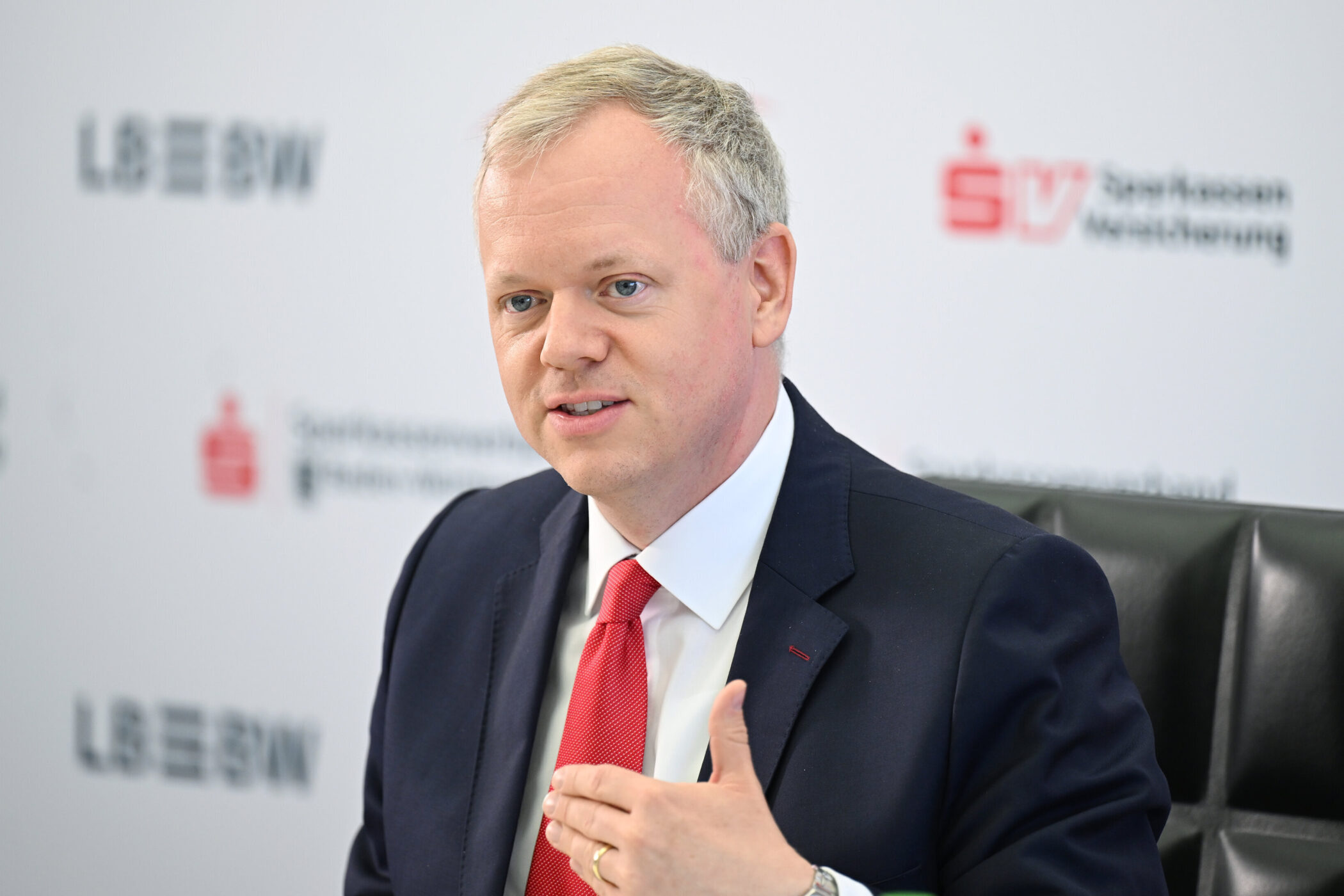 Person in Anzug und roter Krawatte, spricht gestikulierend. Hintergrund zeigt Logos.
