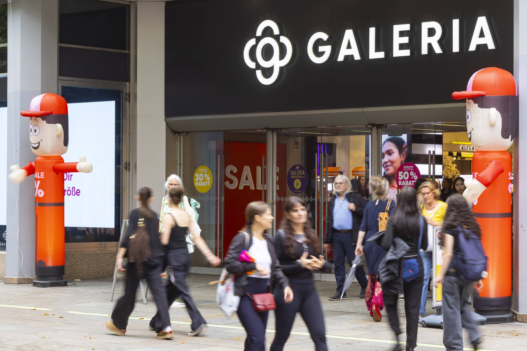 Personen betreten und verlassen ein Geschäft mit "Galeria"-Schild. Luftballonfiguren.