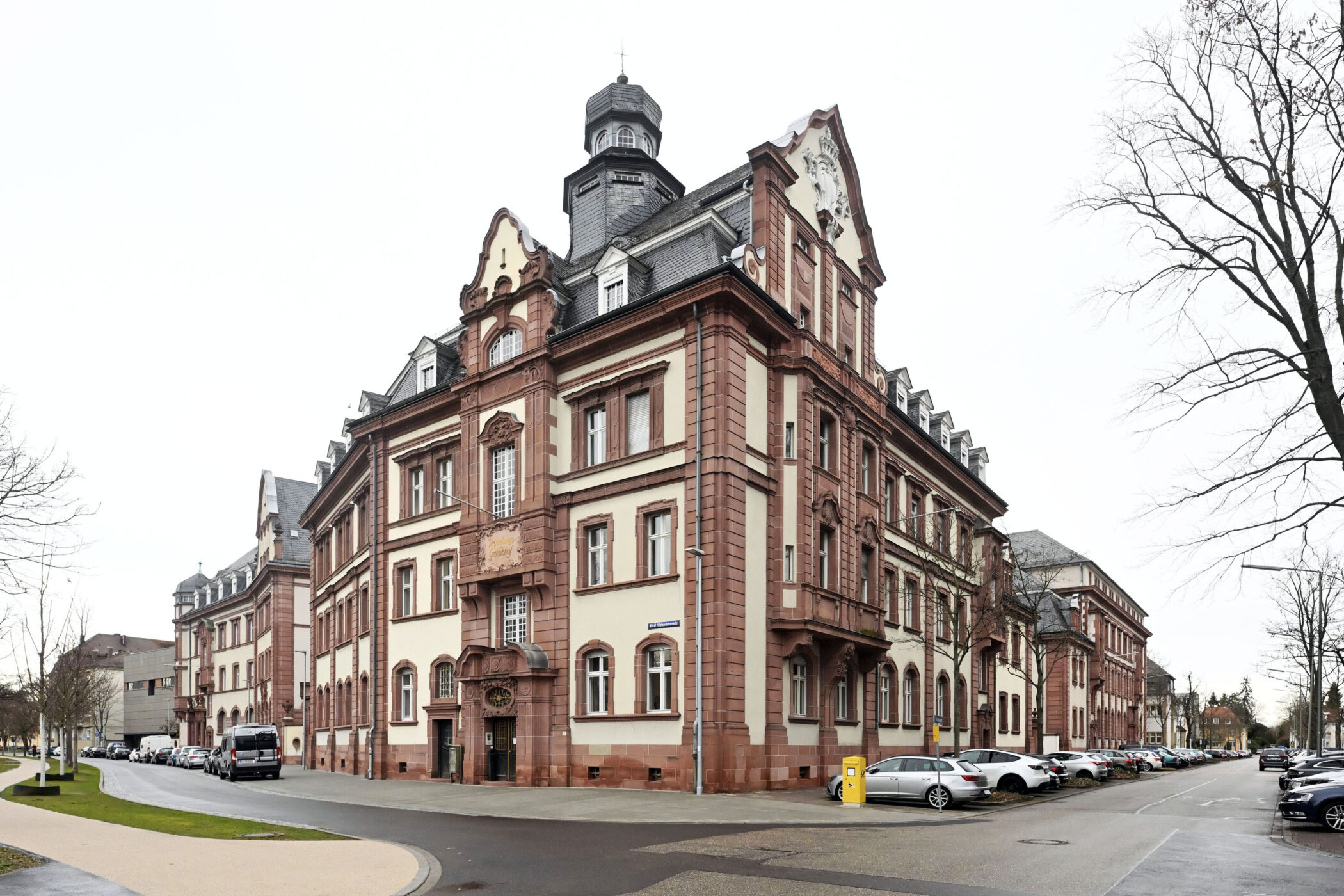 Historisches Gebäude mit rotem Sandstein und Turm, parkende Autos, kahler Baum.