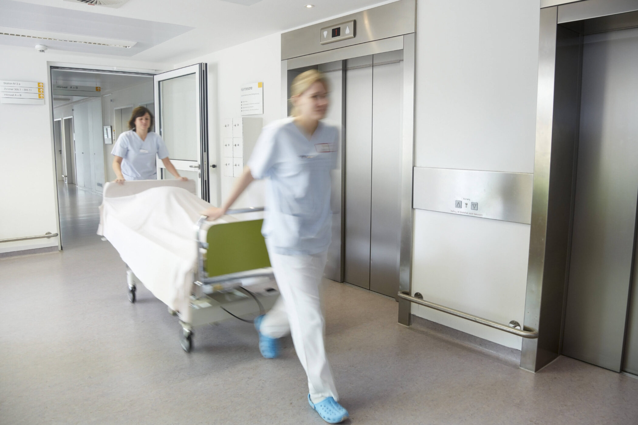Zwei Personen schieben ein Bett durch einen Krankenhausflur.