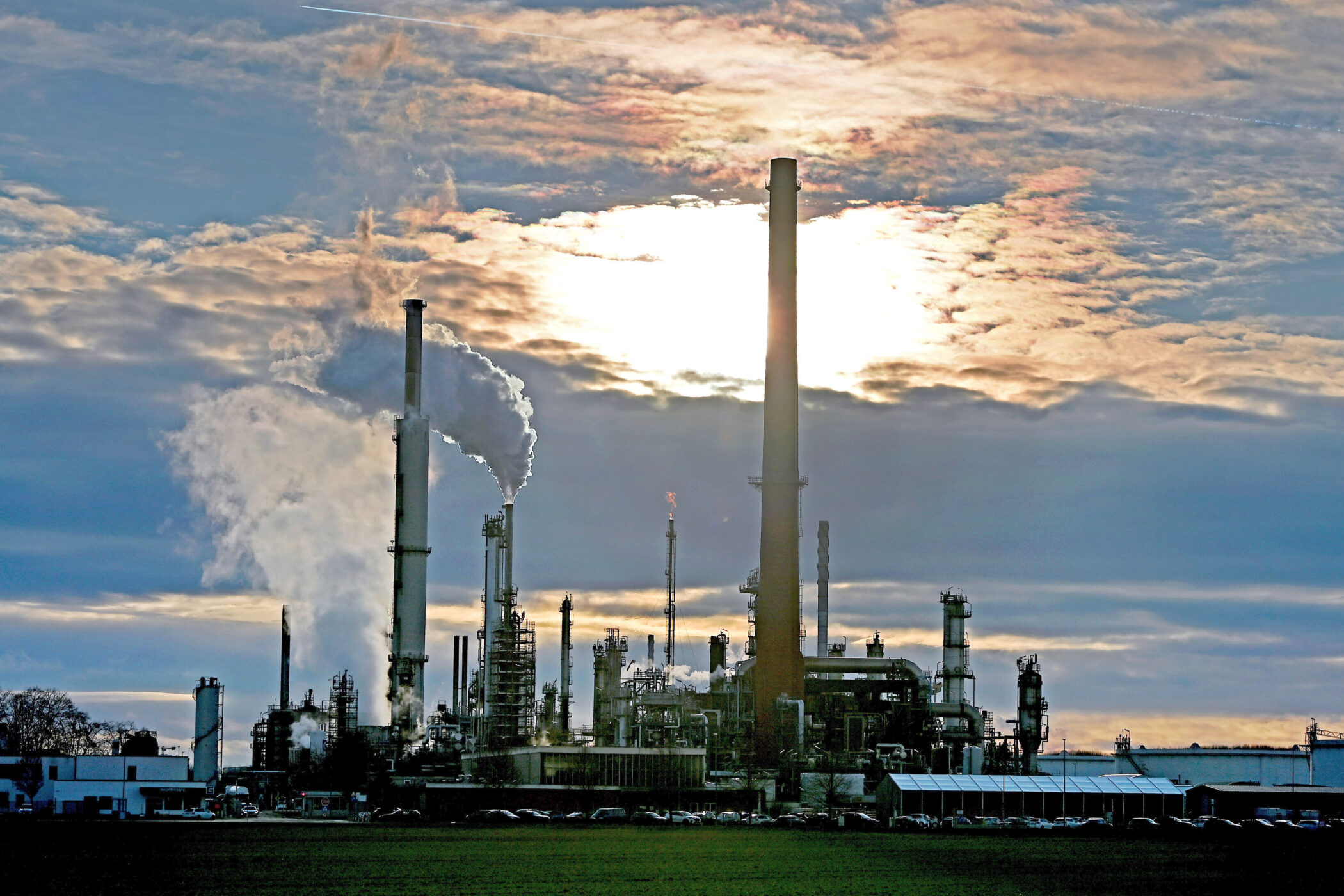 Industrieanlage mit Schornsteinen, Qualm und wolkigem Himmel im Hintergrund.