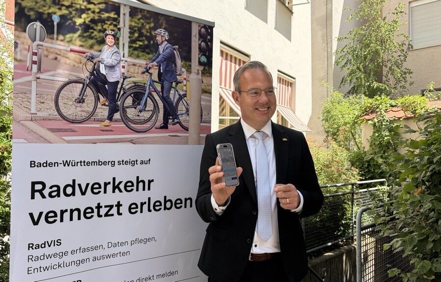 Mann im Anzug zeigt ein Smartphone. Plakat: „Radverkehr vernetzt erleben“.