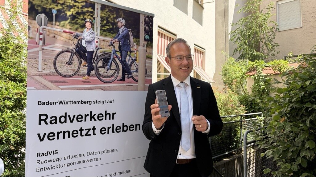 Mann im Anzug zeigt ein Smartphone. Plakat: „Radverkehr vernetzt erleben“.