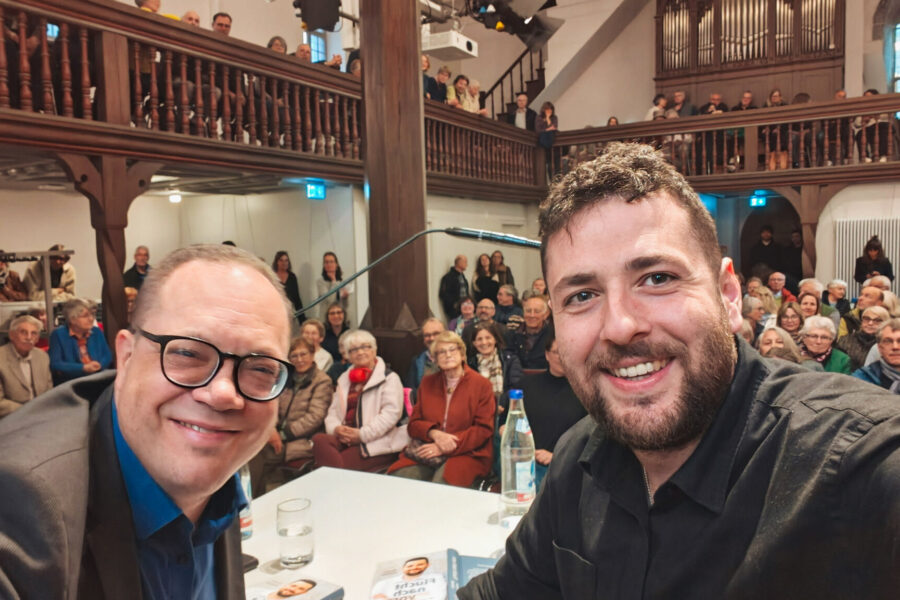 Zwei Männer lächeln in die Kamera, Publikum im Hintergrund in einem Saal.