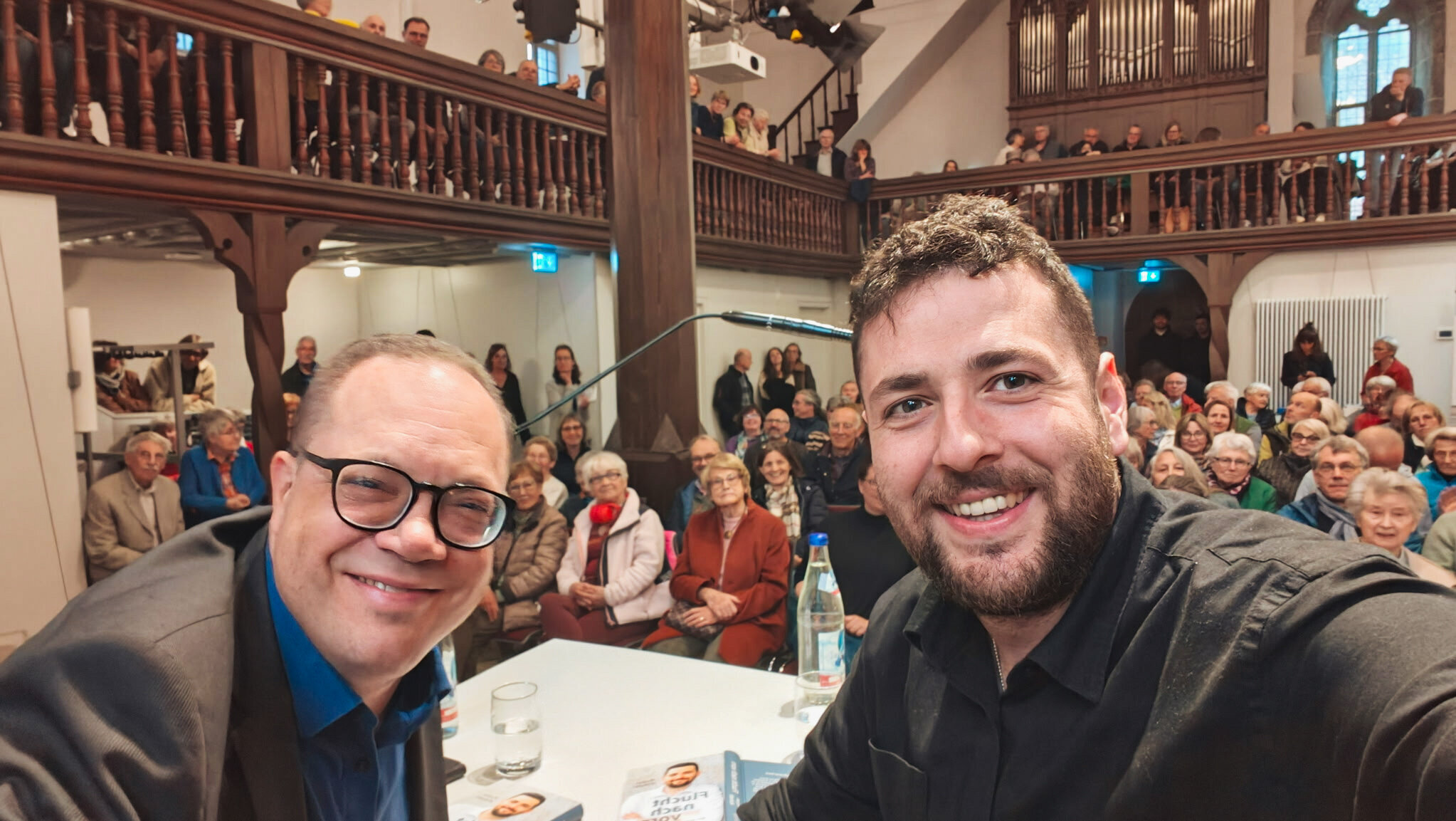 Zwei Männer lächeln in die Kamera, Publikum im Hintergrund in einem Saal.