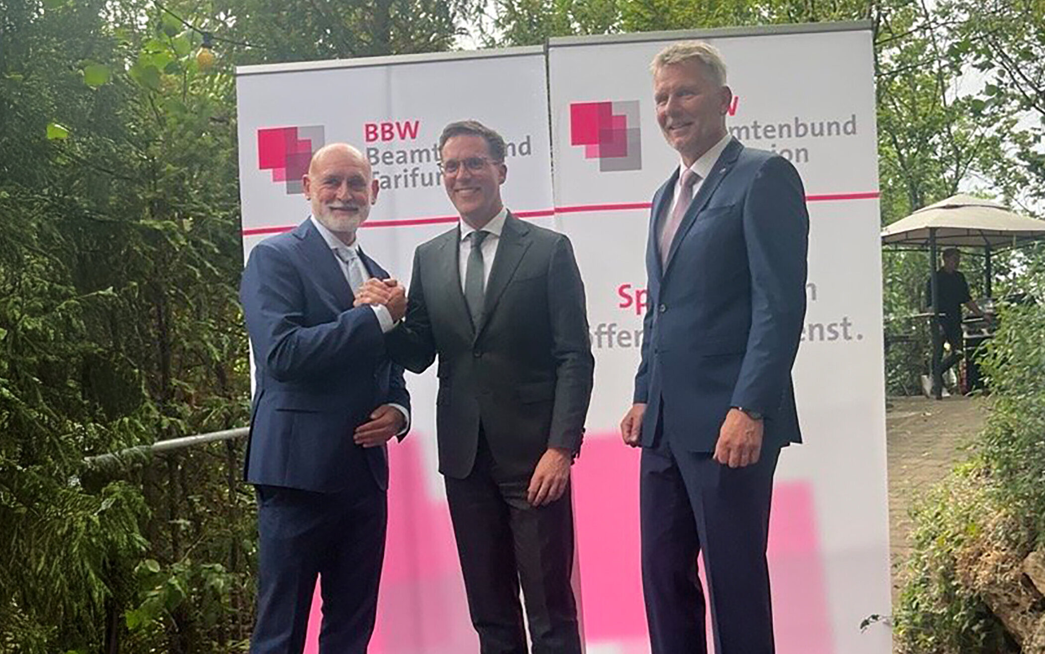 Drei Männer in Anzügen stehen vor BBW-Tafeln im Freien.