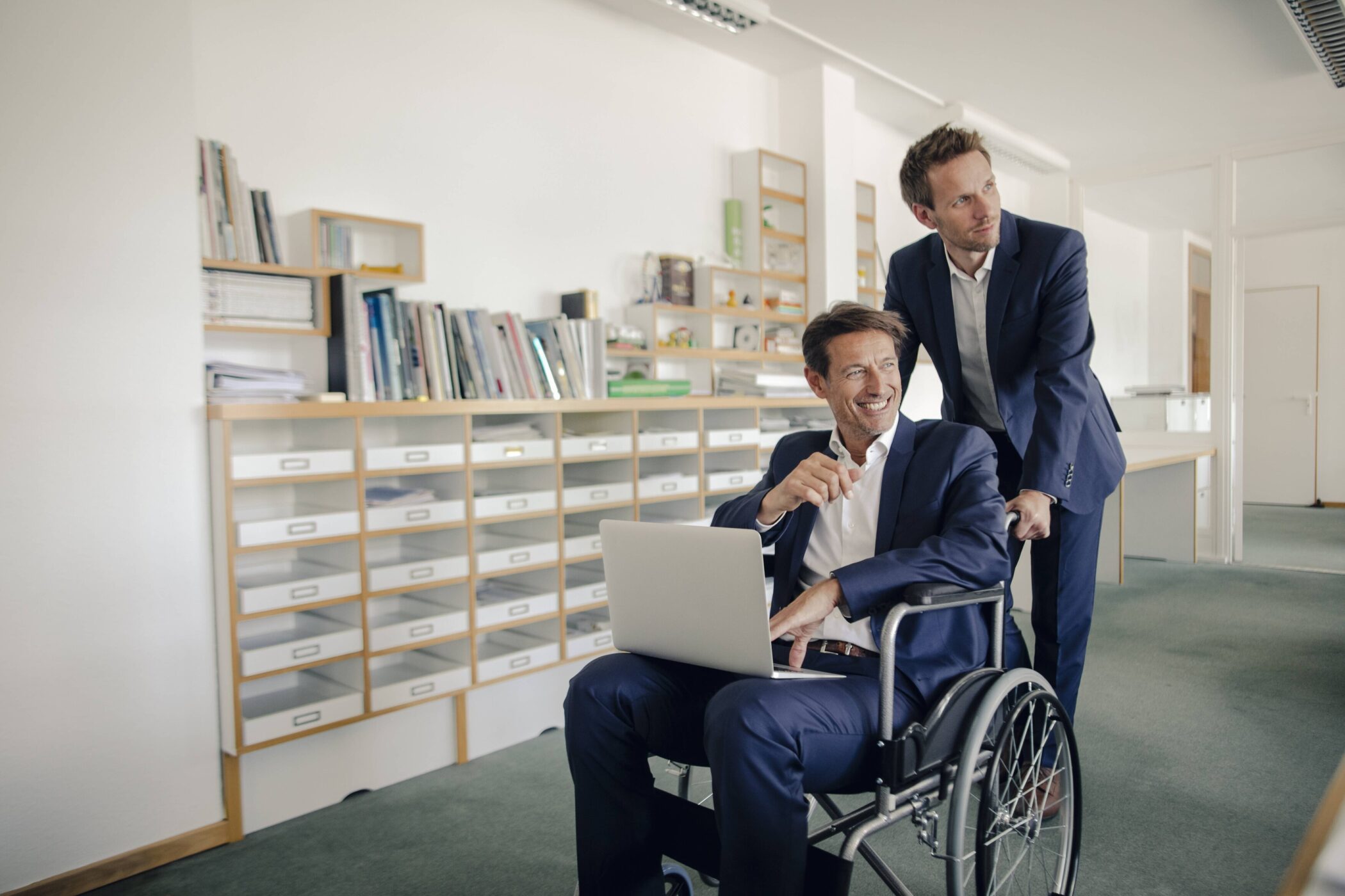 Zwei Männer im Anzug, einer im Rollstuhl mit Laptop, im Büro.