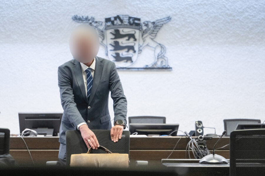Person in Anzug steht an Tisch, zwei Computer, Mikrofon, Wappen im Hintergrund.